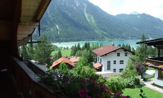 A picturesque mountain landscape with green forests and a clear lake. In the foreground, there is a blooming balcony with colorful flowers.