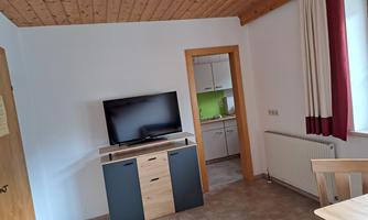 A simply furnished living area with a television and a dining table. In the background, there is an open kitchen.