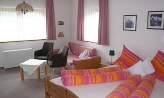 A cozy room with a double bed and colorful pillows. There is a small seating area with two chairs and bright windows.