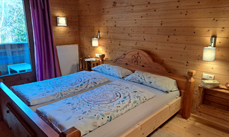 A cozy wooden bedroom with a double bed and colorful blankets. Daylight streams through a window, creating a warm, inviting atmosphere.