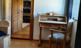 A cozy room with a wooden table and a chair. In the background, there is an open door and a picture on the wall.