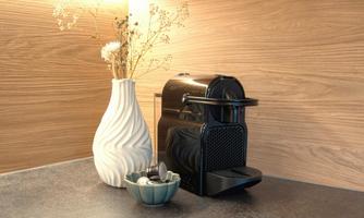 A modern coffee machine sits on a dark countertop. Next to it are a vase with dried flowers and a bowl with coffee capsules.