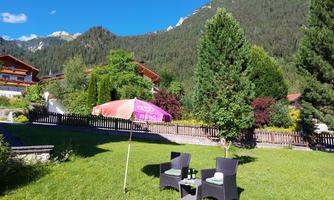 A cozy seating area with two chairs and a table on a green meadow. In the background, trees and mountains can be seen.