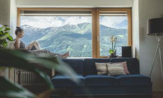 A cozy living room with a blue sofa and large windows. Outside are the snow-covered mountains and a woman sitting relaxed with a book.