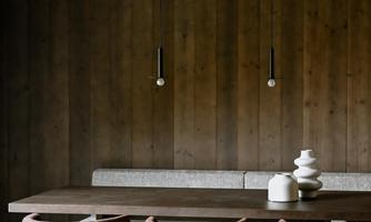 A modern wooden dining table with several chairs is in a cozy room. The walls are clad in dark wood, and minimalist lamps hang from the ceiling.