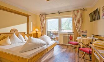 A cozy room with a large bed, bright walls, and wooden details. The room has windows with a view of the mountains and a small seating area.