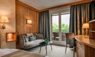 A cozy hotel room with wooden walls and a large window. There is a sofa, a table, and a desk, as well as a balcony with a view of the greenery.