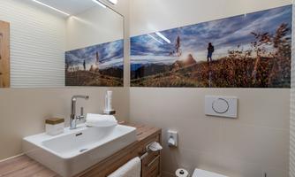 A modern bathroom with a sleek sink and a wooden shelf. On the wall, there is a large panorama picture of a scenic landscape.