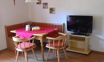 A cozy dining area with a table, chairs, and a bench. In the background, there is a television and some decorative pictures.