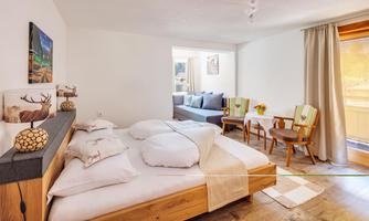 A cozy bedroom with a large bed and light wooden furniture. There is a seating area with two chairs and plenty of daylight.