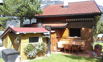A cozy, traditional wooden house with a red roof. In the foreground, there are garden furniture and flowers on the green lawn.