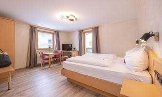 A modern hotel room with a double bed, a table, and chairs. Large windows provide bright light in the room.