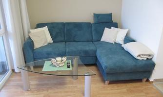 A cozy living room with a blue sofa and a glass table. On the sofa are some cushions and a green tablecloth.