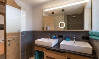 A modern bathroom with two sinks and a large mirror. The decor is minimalist with wooden and gray accents.