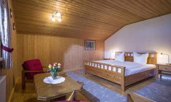 A cozy room with wooden paneling and a large window. There is a double bed, a table, and an armchair, decorated with a flower vase.