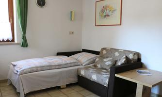 A simple room with a single bed and a sofa. A picture hangs on the wall, and the room has large windows with green curtains.