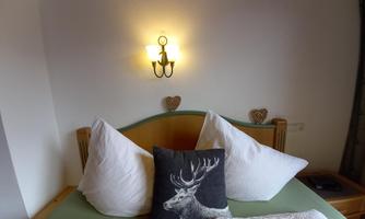 A cozy bedroom with a large bed and decorative cushions. A wall lamp and small wooden hearts give the room a friendly touch.
