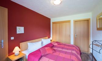 A cozy bedroom with a red accent wall and a double bed. There are bedside lamps and a large wardrobe.