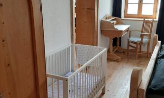 A bright room with wooden floors and white walls. A children's bed stands next to a wooden wardrobe and a desk with a chair.