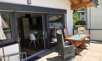 A bright terrace with a wooden table and comfortable chairs. In the background, modern kitchen furniture and a beautiful view of the surroundings can be seen.