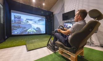 A man is sitting in a racing seat and playing a racing game on a large screen. The room has a modern look with artificial grass and a big screen.