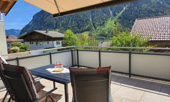 A terrace with a table and chairs offers a beautiful view of green mountains and a blue sky. The surroundings are peaceful and inviting.