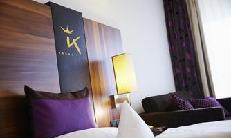 A modern hotel room with an elegant headboard and a cozy sofa. The colors are in shades of purple and wood tones.