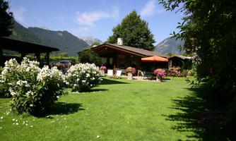 A rustic house in the greenery with blooming flowers and a well-kept garden. In the background, mountains and a blue sky can be seen.