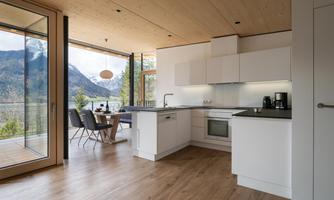 A modern kitchen with bright furniture and wooden floors. Through large windows, there is a view of the mountains and nature.