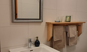 A modern bathroom with a sink, a mirror, and a wooden shelf. Towels and decorative elements can be seen.