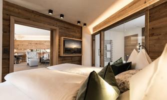 A modern bedroom with two single beds and elegant cushions. The walls are made of wood, and in the background, a bright living area is visible.