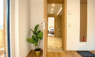 A bright hallway with an open door and a green plant in a gold pot. In the background, a welcoming room can be seen.