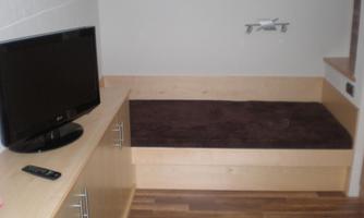 A cozy room with a television and a wooden shelf. The bed has a brown blanket and the floor is made of wood.