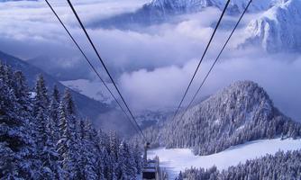 An impressive winter landscape with snow-covered mountains and low-hanging clouds. A cable car runs through the wintry surroundings.