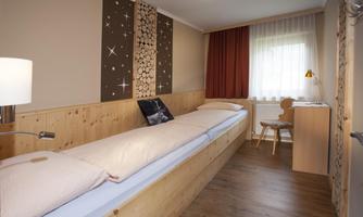 A cozy room with a single bed and wooden decor. Daylight streams through the window, and a desk with a chair complements the furnishings.