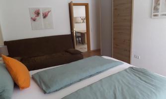 A cozy bedroom with a double bed and elegant pillows. In the background, a sofa and an open room can be seen.