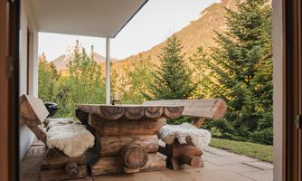 A cozy wooden seating area with fluffy cushions, surrounded by green trees. In the background, gentle mountains can be seen.