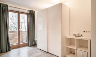 A modern room with a white wardrobe and an open shelf. Large windows with curtains let in a lot of light.