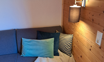A cozy sofa with colorful cushions and a blanket. On the wall, there is a modern lamp and wood paneling.