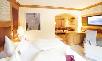 A cozy hotel room with a large bed, soft lighting, and modern decor. A separate living area with a red sofa set and a television is also available.