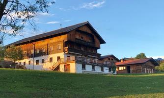 A traditional wooden house in Alpine style, surrounded by green grass. The gentle evening sun illuminates the facade and the sky.