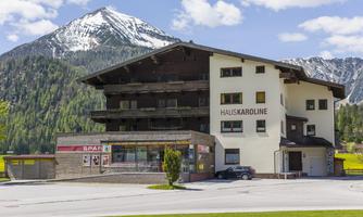 A modern building called Haus Karoline, surrounded by green meadows and majestic mountains. The sky is clear and blue with a few clouds.