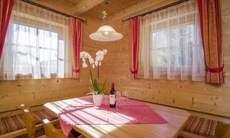 A cozy dining area in a wooden porch with a large table. On the table are a flower and a bottle of wine.