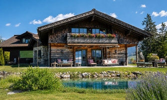 A charming wooden house with a large balcony and colorful flowers. In the foreground, there is a small pond and green grass.
