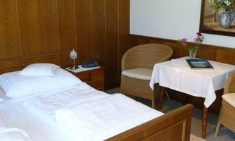 A cozy bedroom with a wooden bed and white bedcovers. Next to the bed is a small table with a flower vase and two chairs.