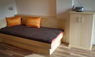 A cozy room with a bed and orange cushions. Next to the bed is a wardrobe and a radio.