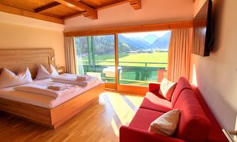 A cozy hotel room with a large bed and a red sofa. The view through the windows shows a picturesque landscape with mountains and meadows.