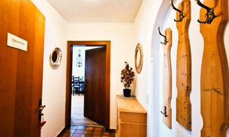A bright hallway with wooden panels and coat hooks. In the background, another room with a table and chairs is visible.