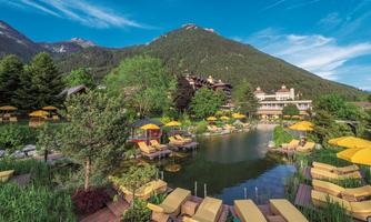 A beautiful wellness area with a pond and loungers. Surrounded by green trees and mountains under a clear sky.