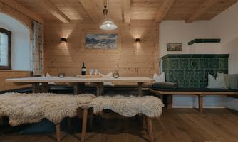 A cozy dining area made of wood with a long table and benches. Paintings hang on the wall, and a traditional stove complements the alpine decor.